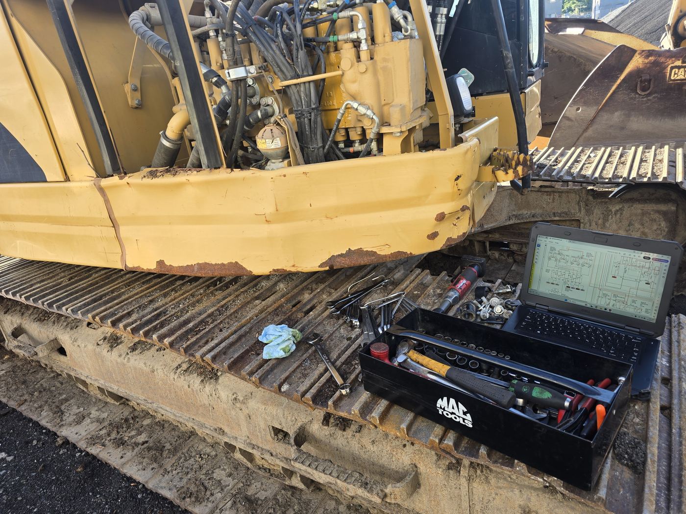 Laptop showing a hydraulic schematic beside a tool tray on the track of a CAT excavator during field diagnostics